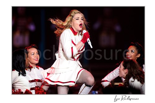 Horizontal color image of Madonna singing into a microphone in a white and red stage outfit with dancers around her during a theatrical live performance