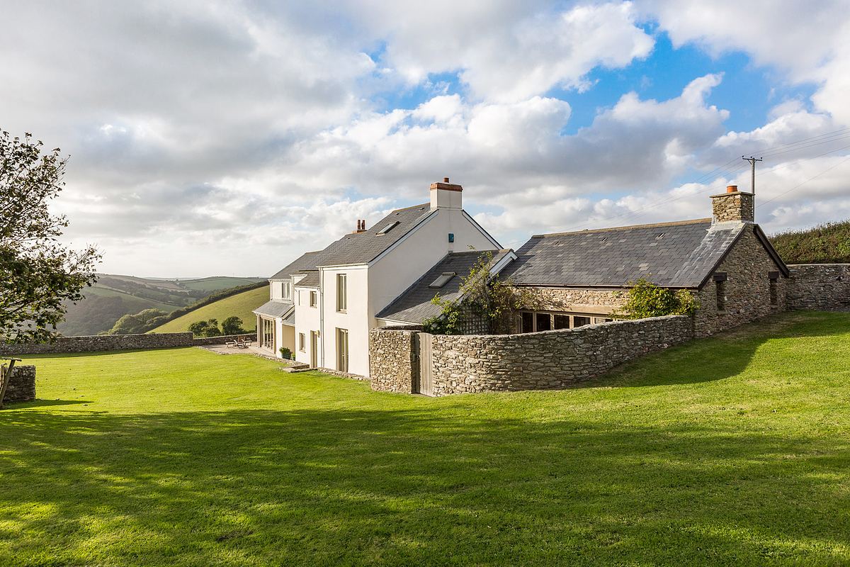 A white countryside house with multiple chimneys, captured by an architecture photographer in Cornwall, stands gracefully surrounded by a grassy lawn and stone wall under a partly cloudy sky.