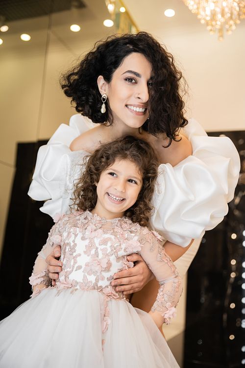gorgeous middle eastern bride with brunette hair standing in white wedding dress with puff sleeves and ruffles behind cute daughter and smiling together in bridal store, looking at camera