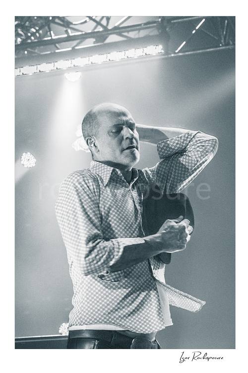 Vertical black and white image of Gord Downie of The Tragically Hip in an emotional live performance moment, holding his hat to his chest with one hand behind his neck under soft stage light