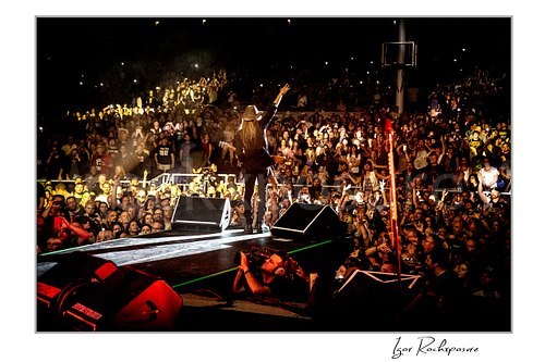 Horizontal color image of C.C. DeVille walking out on stage during a Poison concert toward a packed crowd with raised arm under warm concert lighting