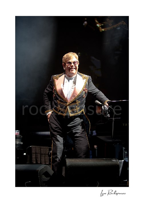 Vertical color image of Elton John standing on stage in an ornate performance suit under dramatic spotlight