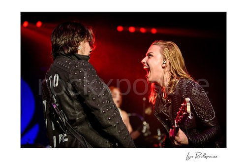 Horizontal color image of Lzzy Hale and Joe Hottinger of Halestorm performing live under vivid red stage light, with Lzzy leaning in toward Hottinger while holding her guitar and smiling fiercely