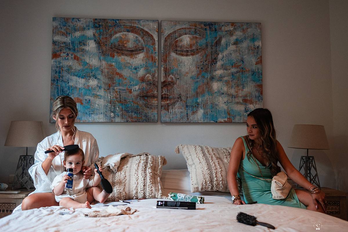 Une mari&eacute;e en peignoir se pr&eacute;pare avec son enfant jouant &agrave; c&ocirc;t&eacute; d'elle et une amie en robe verte assise sur le lit, dans une chambre avec une d&eacute;coration moderne et un grand tableau, photographi&eacute;e par S&eacute;bastien Clavel