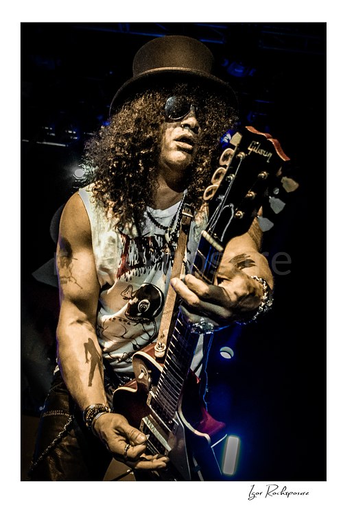 Vertical concert photography of Slash playing electric guitar wearing his signature top hat and sunglasses under dramatic stage lighting