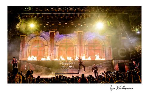 Horizontal color image of Avenged Sevenfold performing on stage in a dramatic full-band wide shot with fire, stage smoke, and a large crowd