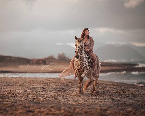 Frau im Palliettenkleid reitet lächelnd auf ihrem Appaloosa am Strand entlang