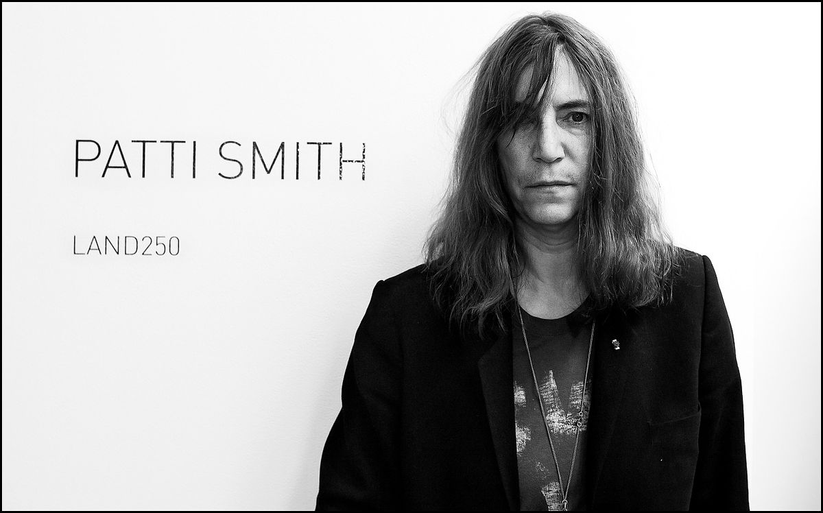 Portrait of singer and musician Patti Smith standing against a art gallery wall with her name printed on it.