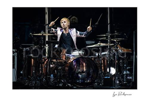 Horizontal color image of Dominic Howard of Muse at the drum kit with both drumsticks raised in a dramatic live performance