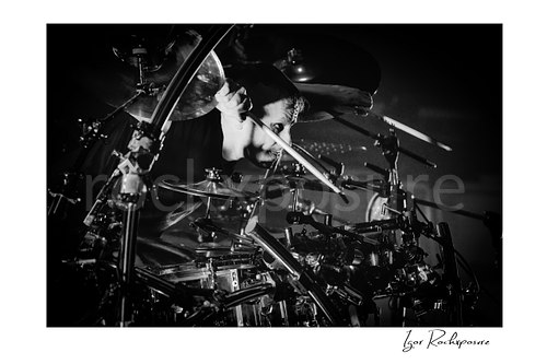 Horizontal black and white image of Korn drummer Ray Luzier leaning forward at the drums with a drumstick pressed along the side of his face during a live performance