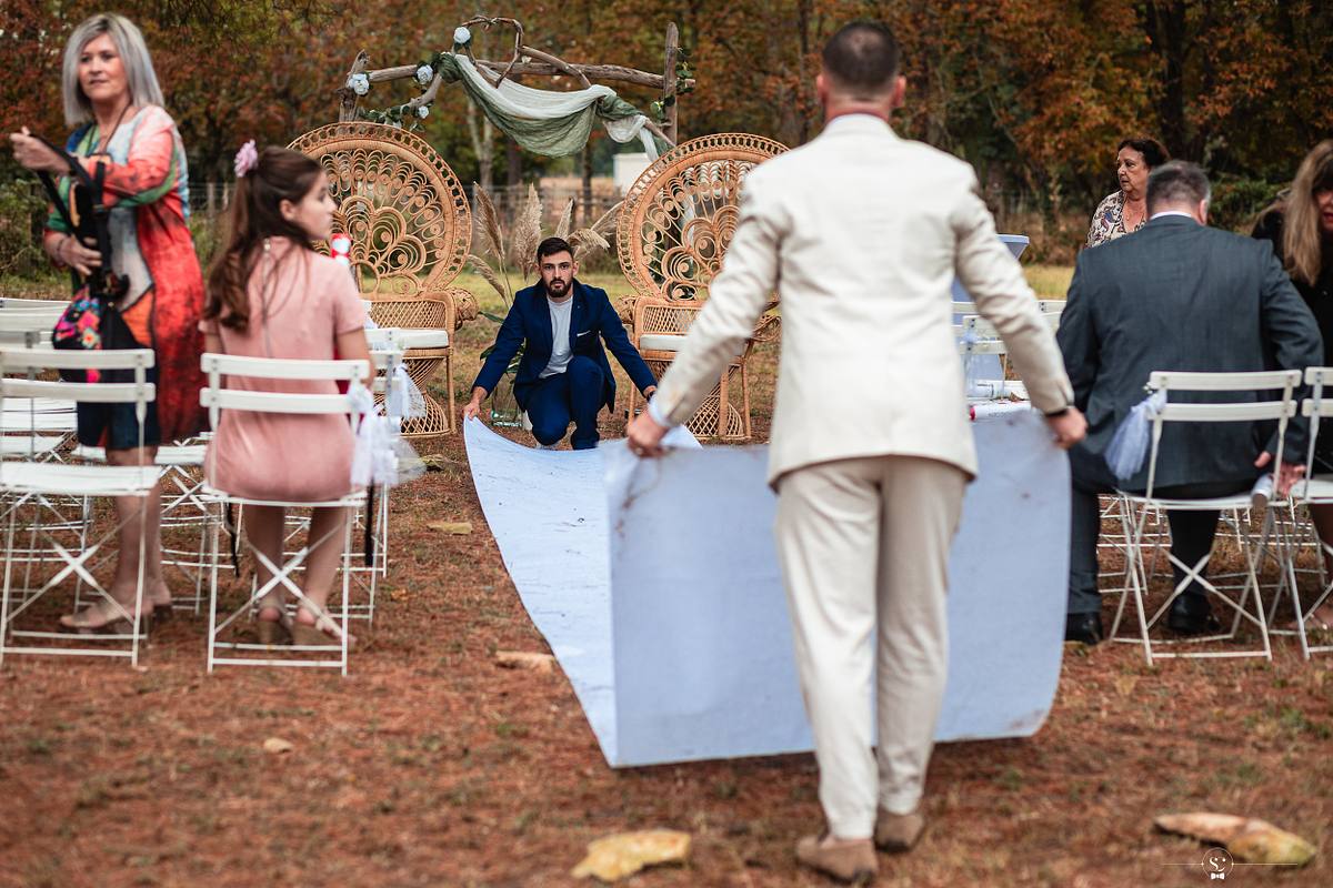 Un homme en tenue de c&eacute;r&eacute;monie s'applique &agrave; d&eacute;rouler un tapis blanc dans la prairie, pr&eacute;parant l'all&eacute;e pour la c&eacute;r&eacute;monie la&iuml;que, entour&eacute; de chaises blanches et de d&eacute;corations rustiques, capturant les pr&eacute;paratifs du mariage champ&ecirc;tre &agrave; N&icirc;mes