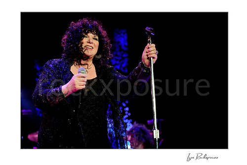 Horizontal color image of Ann Wilson holding a microphone and mic stand during a Heart concert under vivid blue and purple stage light
