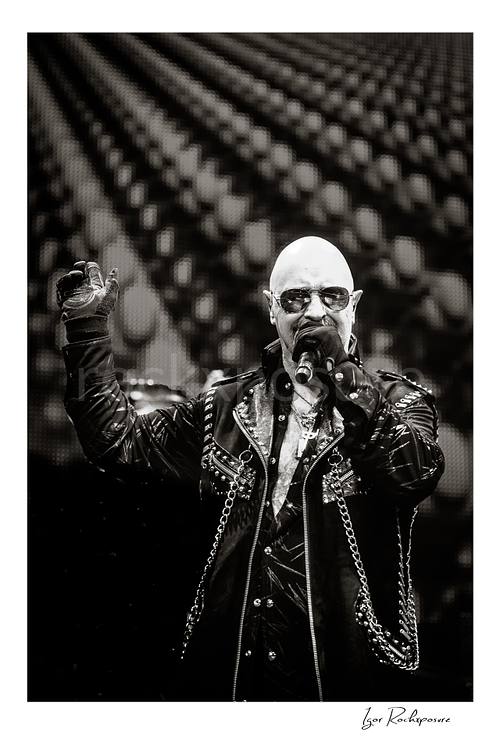 Vertical black and white image of Rob Halford of Judas Priest performing live on stage, singing into a microphone while wearing sunglasses and a studded leather coat