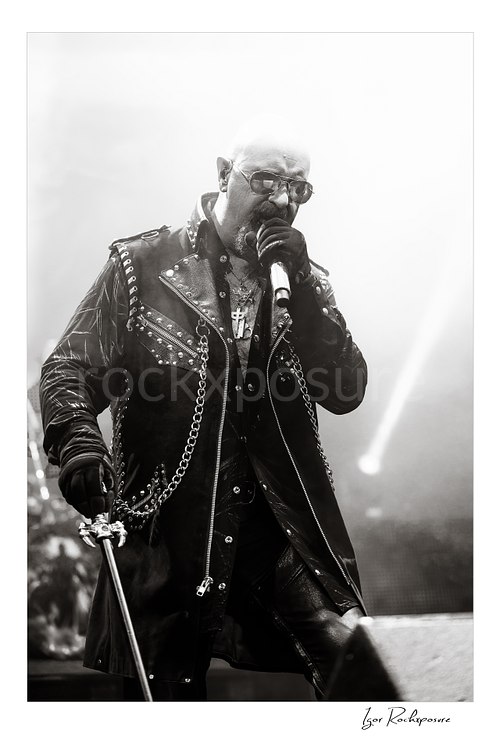 Vertical black and white image of Rob Halford of Judas Priest performing live on stage, singing into a microphone while wearing a studded leather coat and sunglasses