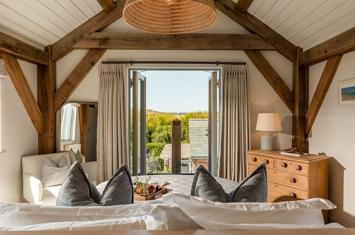 A bright bedroom with exposed wooden beams, a bed with pillows and a tray, a chest of drawers, and French doors opening to an outdoor view—perfect for interiors photography in Cornwall.