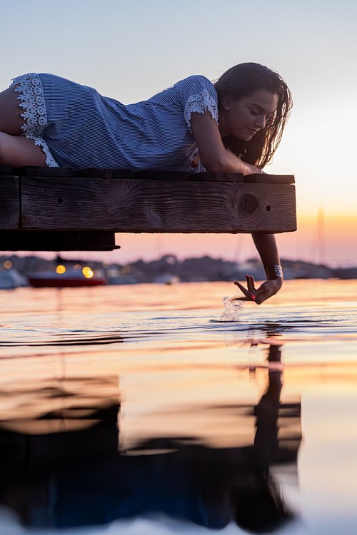 Femme allongée sur un ponton tendant la main vers son reflet dans l’eau
