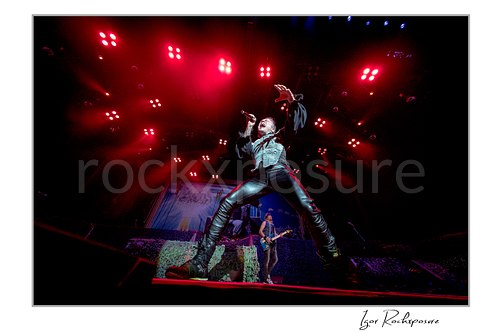 Horizontal color image of Bruce Dickinson of Iron Maiden performing live on stage, singing into a microphone in a dramatic wide stance under red concert lights, with a band member visible in the background.
