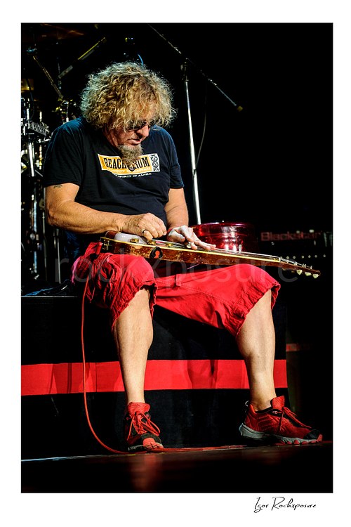 Vertical color image of Sammy Hagar playing guitar onstage in sunglasses under bright backlight and red stage haze