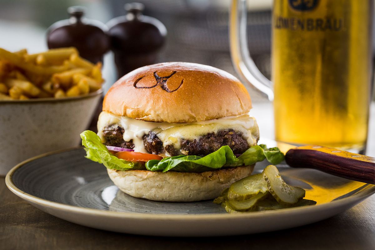 A burger sits on a restaurant table with a glass of beer and some fries blurred out in the background.
