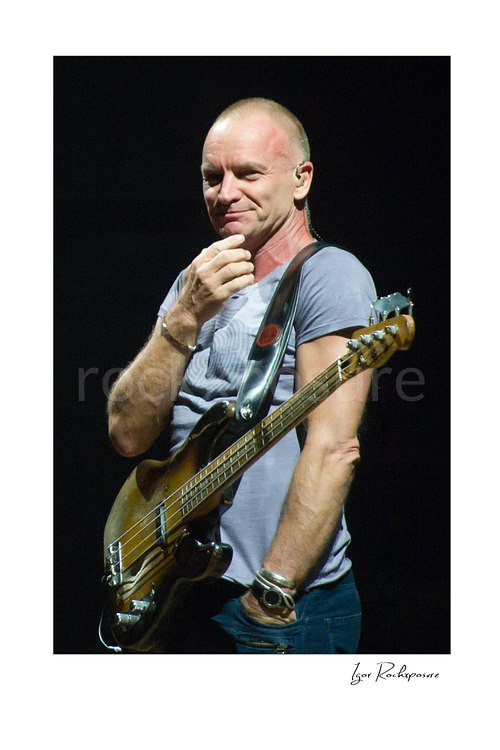 Vertical color image of Sting holding a bass guitar in a quiet live performance portrait under soft stage light