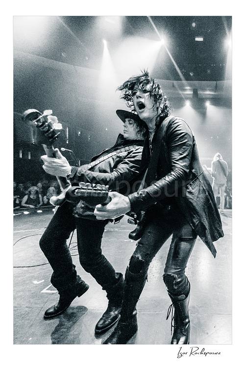 Vertical black and white image of Tom Keifer and Billy Mercer performing live on stage in a dramatic close concert moment, leaning into each other with guitars under bright overhead lights