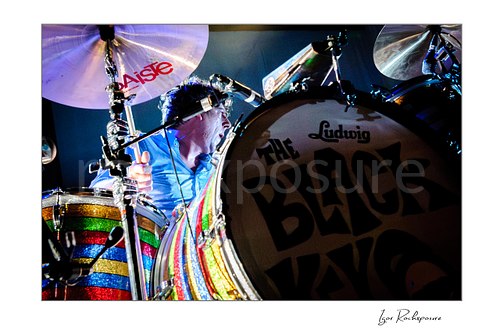 Horizontal color image of Patrick Carney of The Black Keys playing drums behind a rainbow-striped kit with the band logo prominent on the bass drum