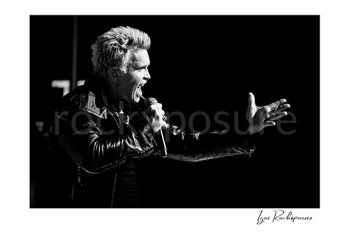 Horizontal black and white image of Billy Idol singing into a microphone with an outstretched hand in a dramatic live performance shot