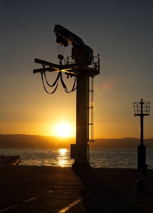 Silhouette eines großen, weißen Hafenkranes auf einem Betonkai bei Sonnenaufgang an der Adria. Die tief stehende Sonne bildet eine leuchtende Korona hinter dem Kran und taucht den Himmel sowie die Wasseroberfläche in ein warmes, goldenes Licht. Im Hintergrund zeichnen sich die sanften Umrisse einer Gebirgskette ab.