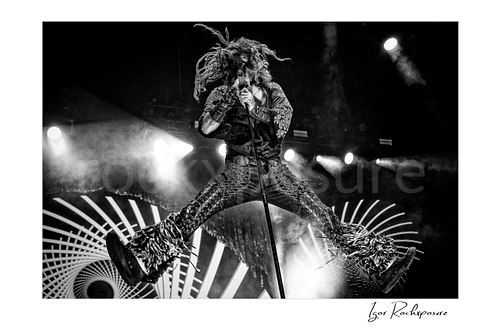 Horizontal black and white image of Rob Zombie performing live on stage in a midair jump with dreadlocks flying, fringed pants, and a microphone stand