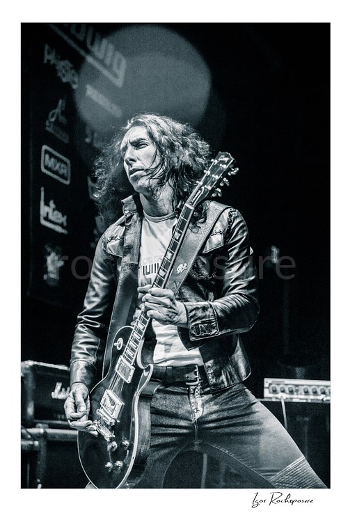 Vertical black and white image of Brent Woods playing guitar on stage wearing a leather jacket in a dramatic performance shot