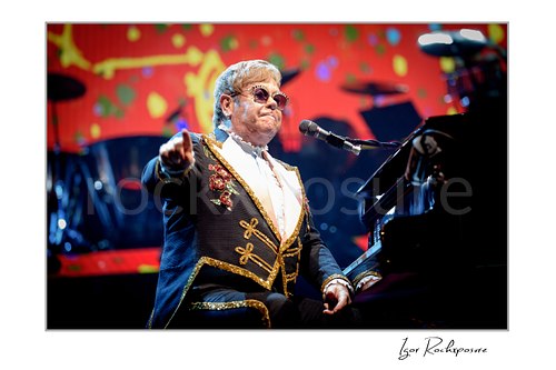 Horizontal color image of Elton John seated at the piano pointing outward against a vivid red stage backdrop