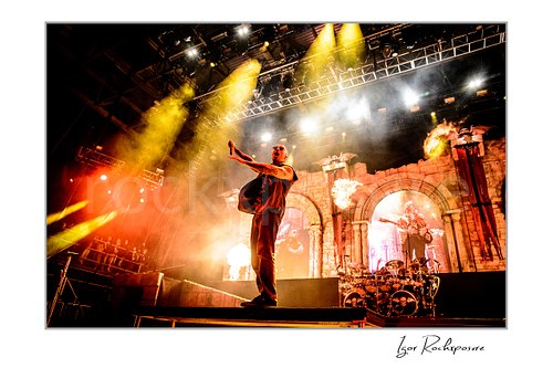 Horizontal color image of M. Shadows of Avenged Sevenfold performing on stage with outstretched arms, fire bursts, and dramatic yellow stage lights