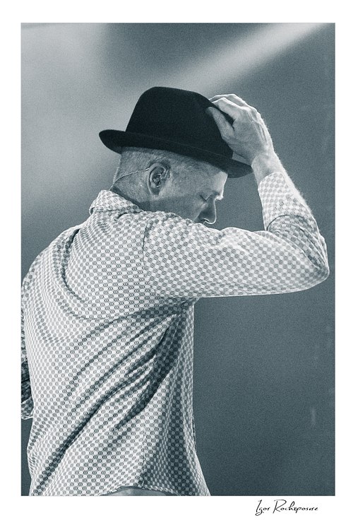 Vertical black and white image of Gord Downie of The Tragically Hip in a quiet live performance moment, touching the brim of his hat under soft stage light