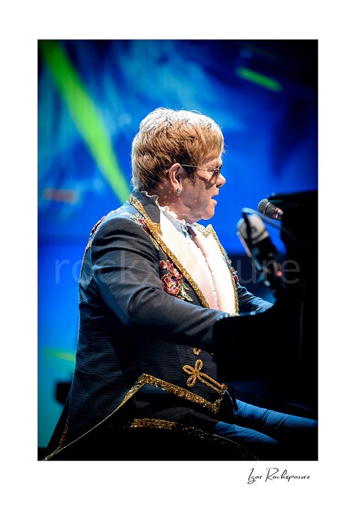 Vertical color image of Elton John seated at the piano in profile under blue stage lighting
