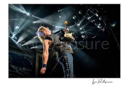 Horizontal color image of Dave Murray of Iron Maiden performing live on stage, leaning back while playing electric guitar under bright white concert lights