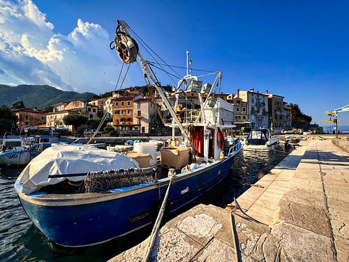 Ein traditionelles blaues Fischerboot mit einem großen Netzkran liegt im malerischen Hafen von Moščenićka Draga in Kroatien an der Kaimauer, im Hintergrund bunte Küstenhäuser unter blauem Himmel.