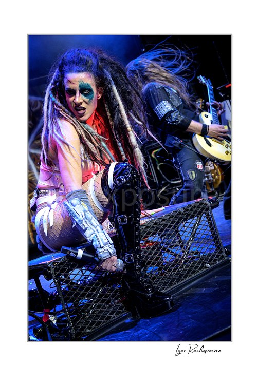 Vertical color image of Calico Cooper of Beasto Blanco in a fierce live performance pose on stage, with dramatic makeup, long dreadlocks, and another guitarist blurred behind her