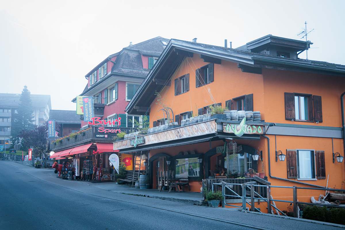 Nebeliger Blick auf die Dorfstrasse von Grindelwald mit der orangefarbenen Fassade der Avocado Bar, dem Sportgesch&auml;ft Buri Sport und Souvenirl&auml;den