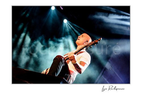 Horizontal color image of Johnny Christ of Avenged Sevenfold playing bass onstage beneath bright concert beams and drifting smoke.