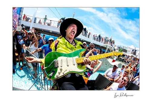 Horizontal color image of Randy Hansen playing a green guitar beside the pool during an outdoor ship deck performance with cheering fans around him