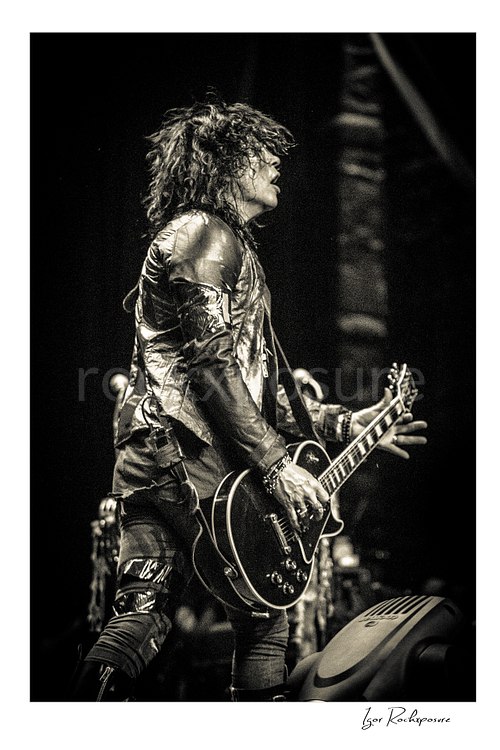 Vertical black and white image of Tom Keifer performing live on stage with a black guitar, captured in side profile with tousled hair and bright stage light behind him