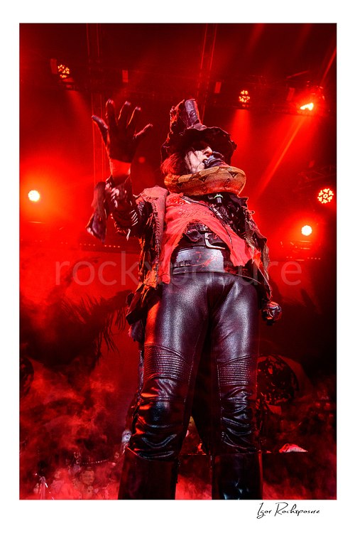 Vertical color image of Alice Cooper performing live on stage under red lighting in a black top hat, red scarf, and black leather outfit with a gloved hand raised