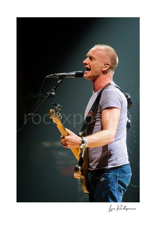 Vertical color image of Sting singing into a microphone while holding a bass guitar during a live performance