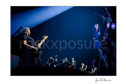 Roger Waters plays black bass at stage left beneath a broad blue beam, with raised hands below and a large projected figure at right.