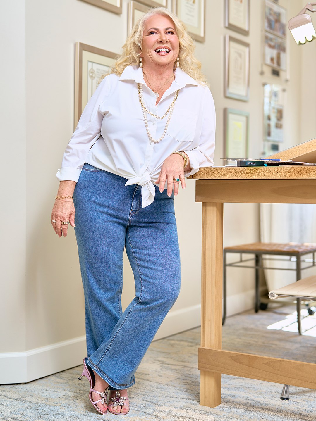 A woman with blonde hair wearing a white blouse and blue jeans stands against a drafting table in her home interior design studio