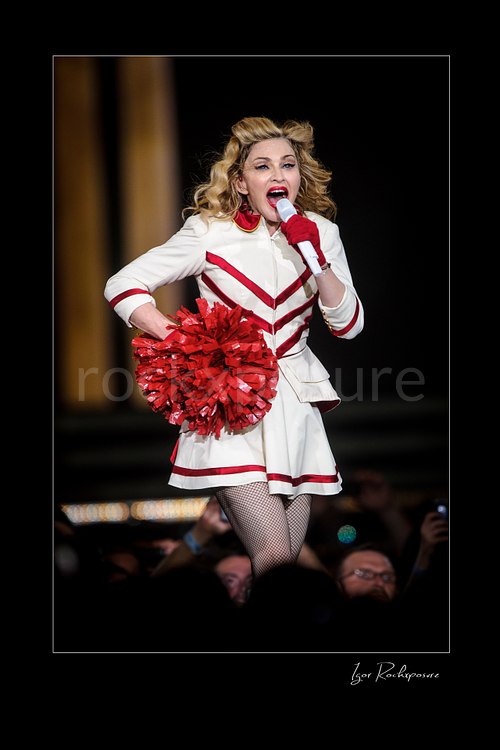 Vertical color image of Madonna singing into a microphone in a white and red stage outfit while holding a bright red pom-pom