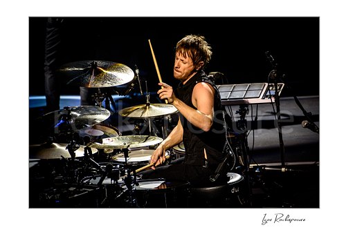 Dominic Howard of Muse Live Concert Print  Horizontal color image of Dominic Howard of Muse playing drums with one stick raised in a focused concert moment against a dark background