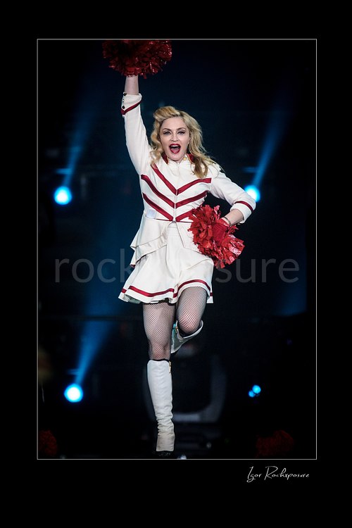 Vertical color image of Madonna performing in a white and red stage outfit with red pom-poms and one arm raised under cool blue concert lights