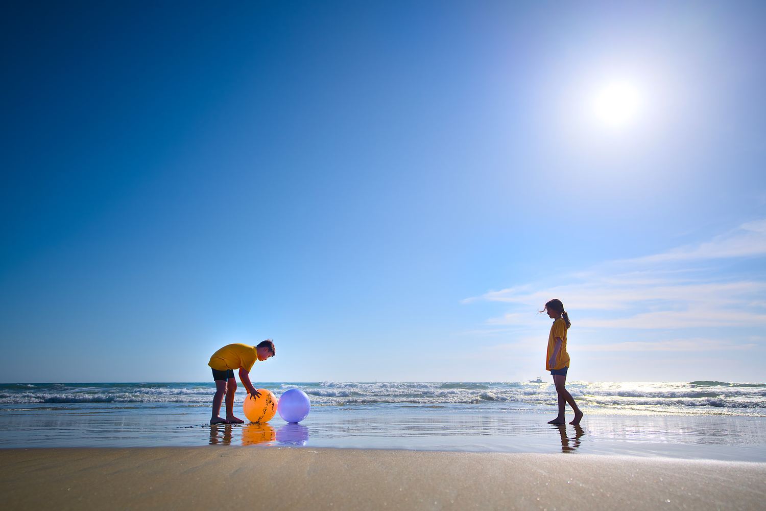 Two children playing with bright beach balls near the shoreline under a clear sky.