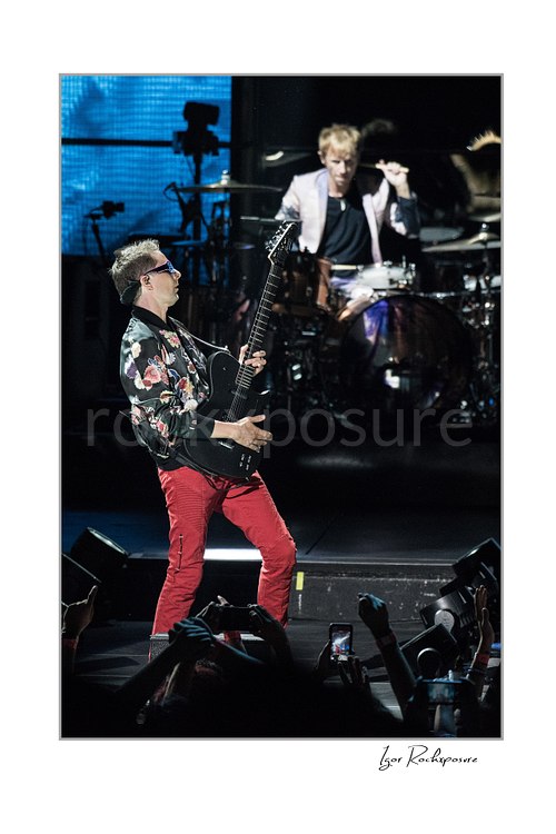 Vertical color image of Matt Bellamy and Dominic Howard of Muse performing live on stage with guitar in the foreground and drums behind